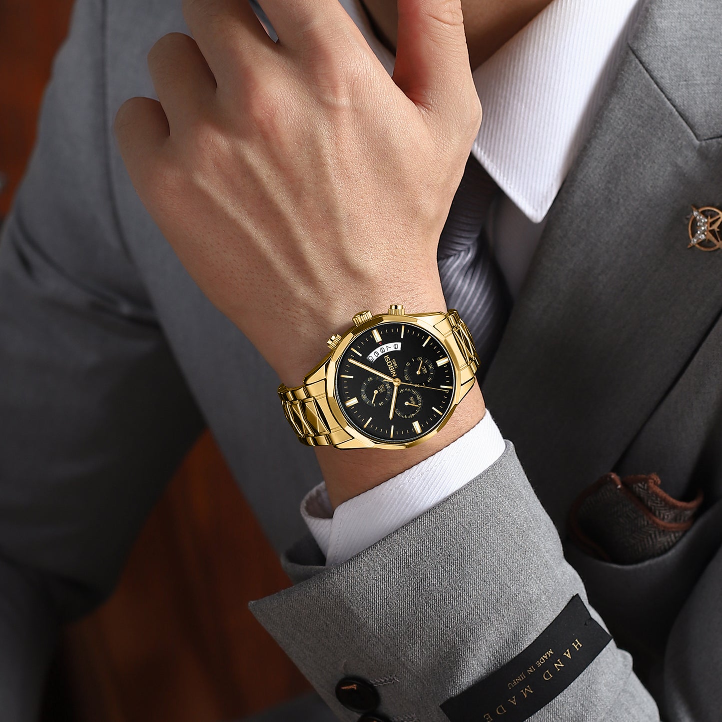 A close-up of a man's wrist wearing a gold-tone metal watch with a black dial, styled with a gray suit jacket, white dress shirt, and striped tie.