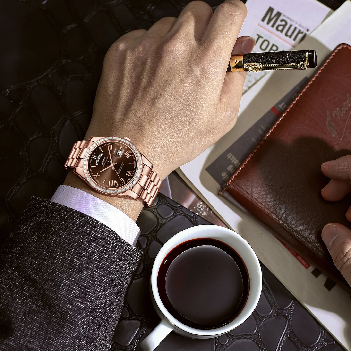 Hand wearing a rose gold watch with a cup of coffee and leather wallet on a textured surface