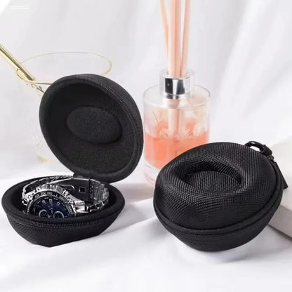 Black travel case with a watch inside, placed on a white surface with a glass and diffuser in the background.